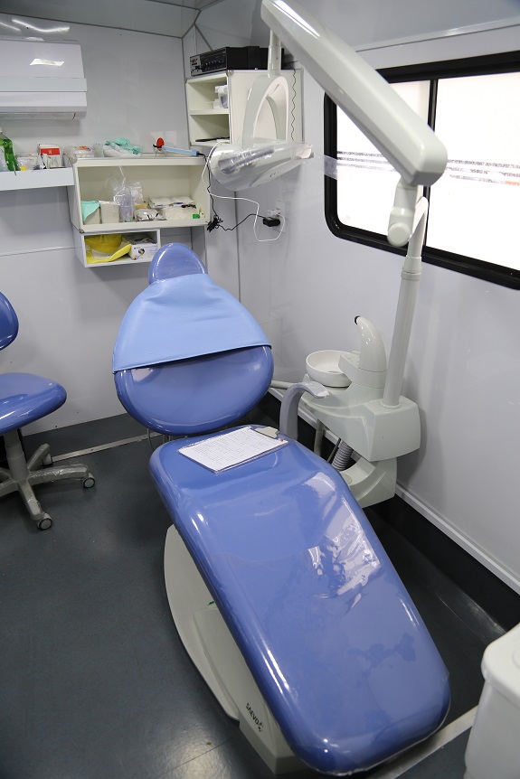 Parte interna do ônibus, equipado com materias para atendimento odontológico. Ao centro, uma cadeira reclinável azul. Ao fundo, nas prateleiras, medicamentos e acessórios para odonto.
